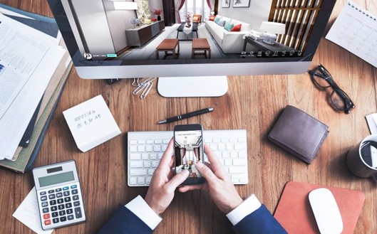 A desk with a computer and a phone both showing a living-room render